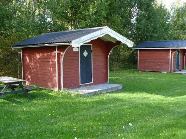 En trästuga omgiven av en välskött gräsmatta på Orbadens camping, med skuggande träd och en fridfull utomhusmiljö.