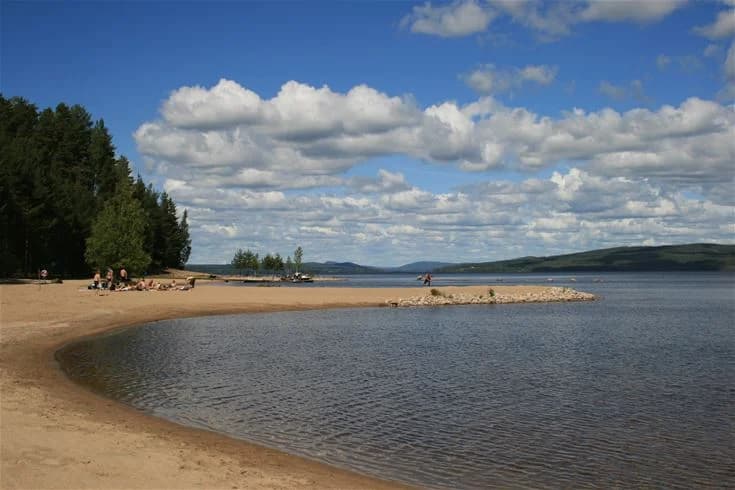 Vy över Orbadens camping med en vacker sjö i förgrunden, omgiven av grönskande stränder och lummiga träd. I bakgrunden syns moln på himlen som speglas i det lugna vattnet.