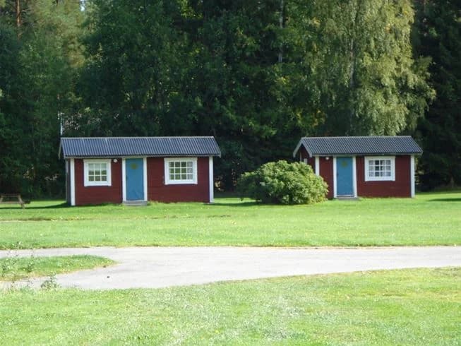 En grön gräsmatta vid Orbadens camping med små stugor och trädgårdsbyggnader omgivna av öppna ytor och träd.