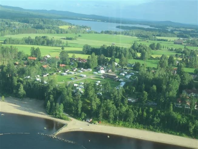 En naturskön vy över Orbadens camping med en glittrande sjö i förgrunden, omgiven av frodiga träd och ett öppet landskap. Kusten sträcker sig längs med vattnet, vilket ger en inbjudande plats för rekreation och avkoppling.