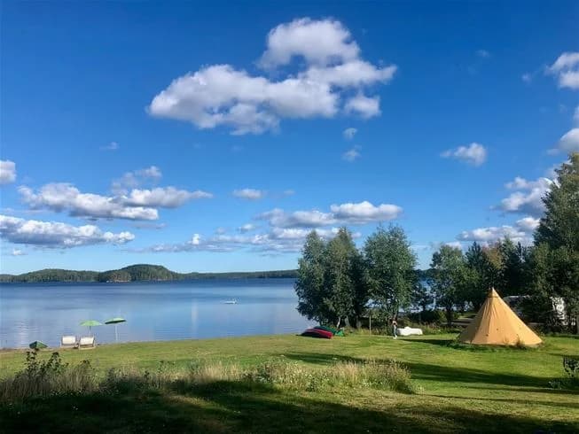 Vy över en campingplats vid en sjö med tält uppställda på en grön gräsmatta vid strandkanten.