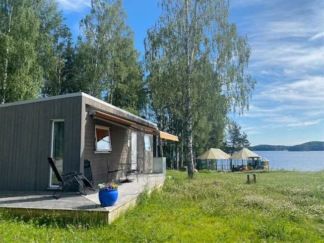 En liten timrad stuga omgiven av trädgårdsbyggnader och skugga, med en mysig och naturnära atmosfär vid Sagaliden.