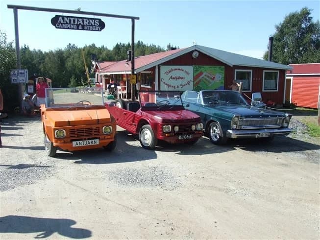En klassisk bil parkerad på Antjärns Camping, omgiven av gröna träd och en grusbelagd parkering. Detaljer som bilens dörr och stötfångare är tydligt synliga.