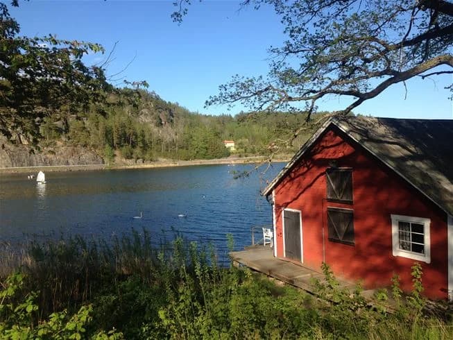 En bild av Grännäs Camping och Stugby med en stuga vid kanten av en sjö. Grönskande vegetation omger vattnet som speglar den klara sommarhimlen.