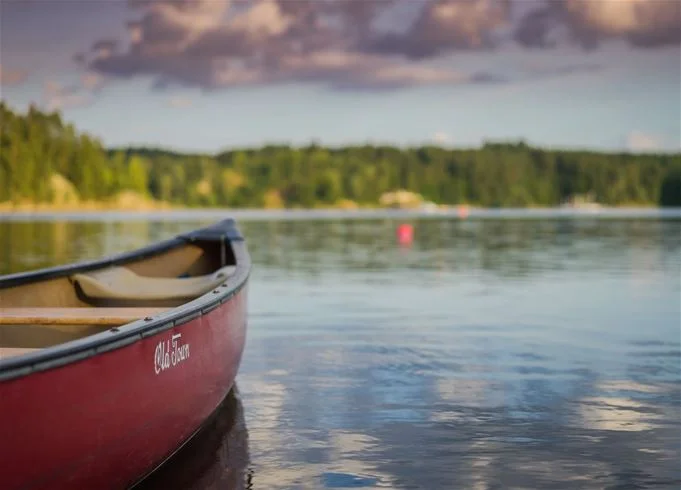 En båt ligger förankrad vid en kaj vid Kalmar camping - Rafshagsudden, omgiven av lugnt vatten som reflekterar träden och den klara himlen.