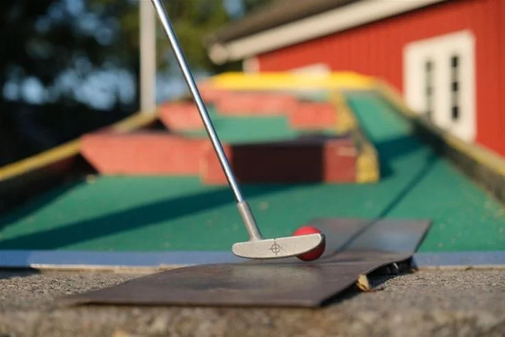 En person spelar minigolf på Gullbrannagården, med fokus på en golfklubba och boll på en minigolfbana.