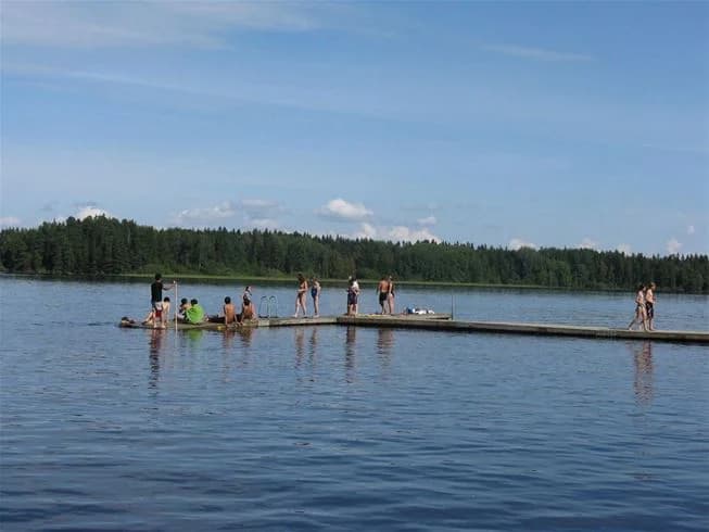 En naturskön utsikt över en sjö med lugnt vatten och moln på himlen, omgiven av gräsbevuxna stränder vid Falkudden camping, café och stugby, en plats för rekreation och avkoppling.
