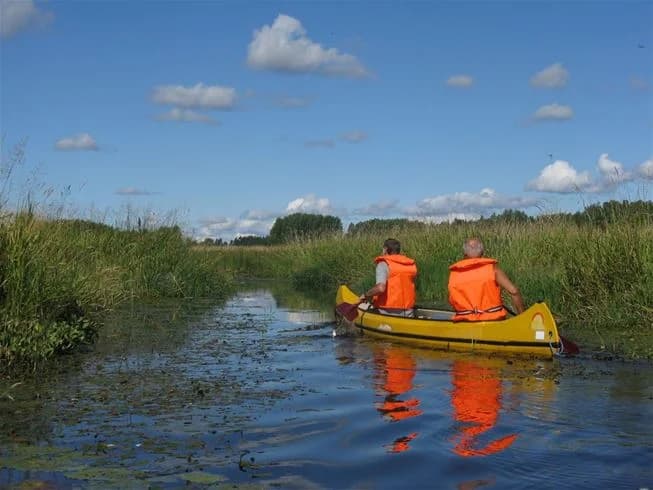 En liten båt ligger förtöjd vid en brygga vid en sjö omgiven av lummig grönska och vass. Det är en solig dag och spegelblanka vattnet bjuder in till rekreation och avkoppling vid Falkudden Camping, Café och Stugby.