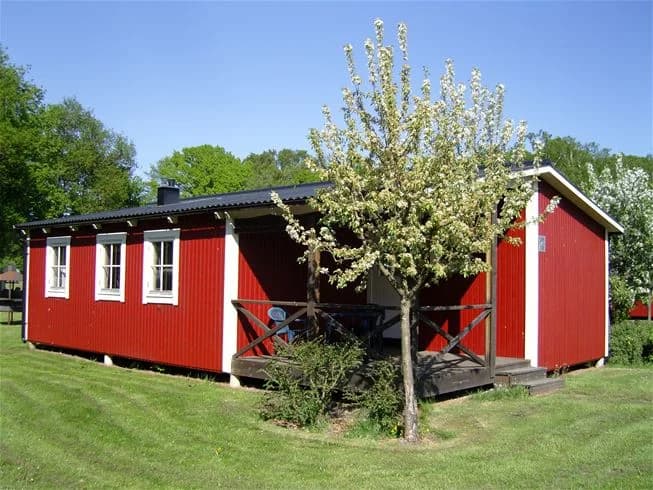 En lantlig stuga omgiven av en grönskande trädgård och gräsmatta, med skugga från närliggande träd och enkla uthus i bakgrunden.