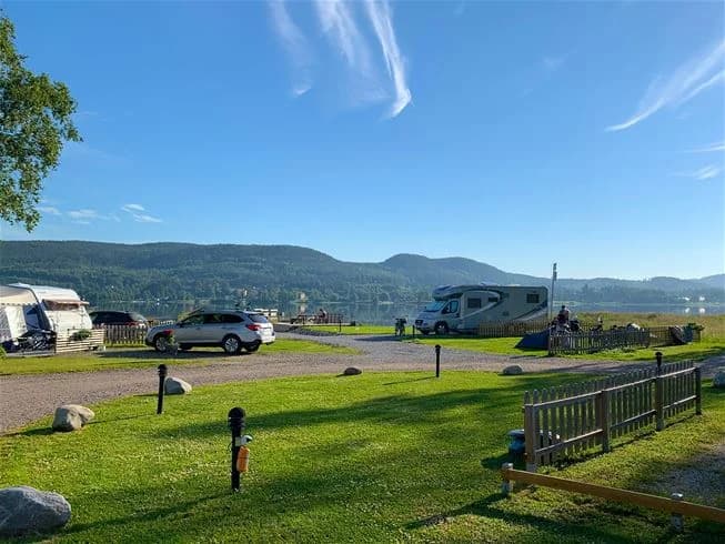 En husvagn står parkerad vid en campingplats med utsikt över en sjö. Ett picknickbord och bänkar finns i förgrunden, omgivet av gröna träd och en klarblå himmel med vita moln.