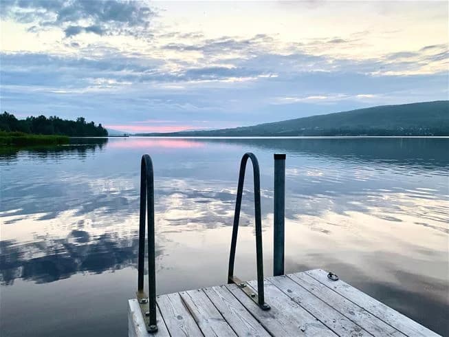En solig sommardag vid Överhörnäs camping, med blått vatten och en klar horisont. Sjön speglar omgivningen, och vid vattnet finns en brygga som sträcker sig ut över den lugna ytan.