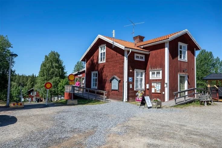 En mysig stuga med träfasad och uppfart syns omgiven av en grönskande trädgård. Framför stugan finns utemöbler och en motorcykel.