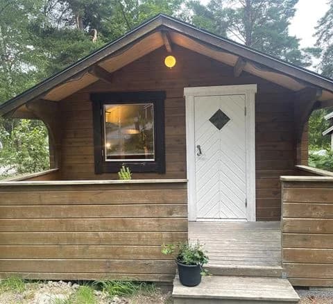 En timmerstuga med trädörr, omgiven av gröna träd och buskar, på Waxholms camping.