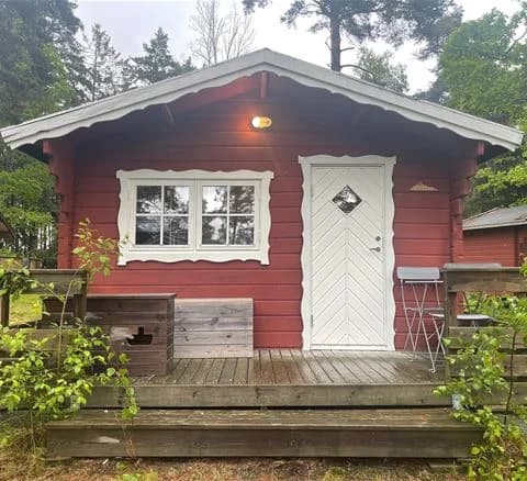 En trästuga med dörr och fönster, omgiven av grönska och träd vid Waxholms camping.