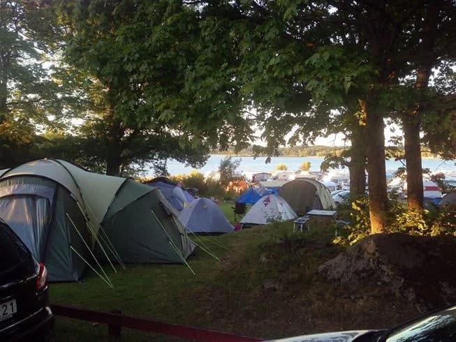 En familjebil parkerad vid ett tält på Lövekulle camping, omgivet av campingutrustning och skugga från ett tarpaulin, perfekt för en avkopplande campingupplevelse.