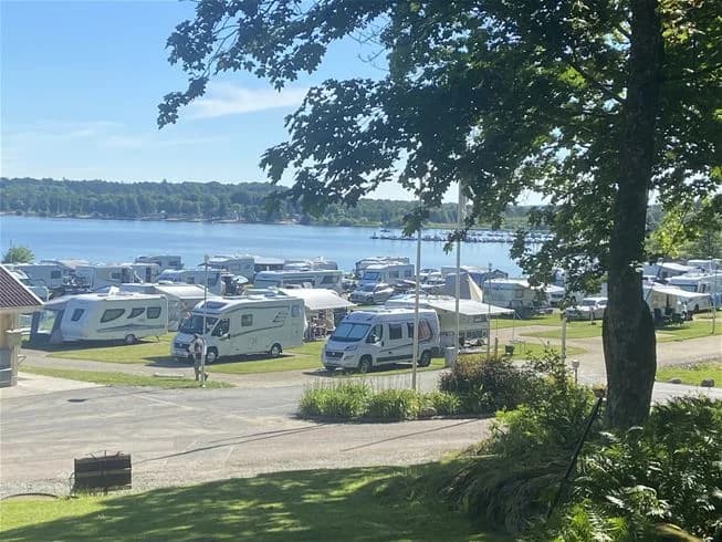 En solig dag vid Lövekulle Camping med husbilar och husvagnar parkerade nära stranden. Flera båtar ligger förtöjda vid kusten och en småbåtshamn syns i bakgrunden.