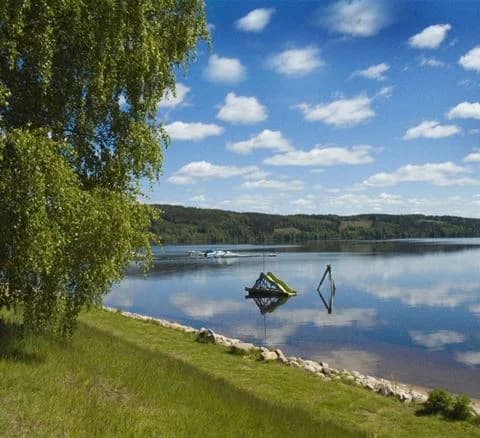 En lugn sjö omgiven av frodig grönska vid Torsby camping, med blå himmel och reflekterande vatten.