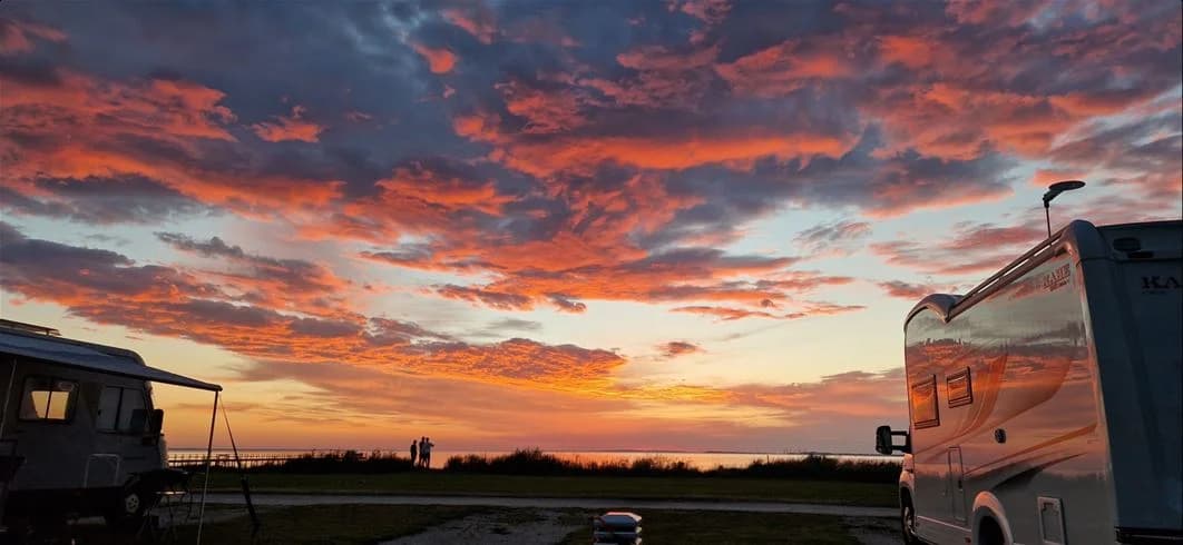 Solnedgång vid Barsebäckstrands camping med en himmel som glöder i orange och rosa toner. Siluetter av moln sträcker sig över horisonten medan en husbil ses i förgrunden, vilket ger en känsla av friluftsliv.