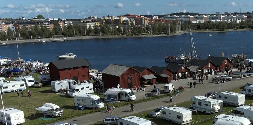 Bild av Västra Kajen Camping: En hamnområdesmiljö med båtar förtöjda vid kajen, en lastbil synlig i bakgrunden, samt ett par tält uppsatta nära vattnet. Morgonsolens ljus speglas i vattnet och skapar en inbjudande atmosfär vid campingen.