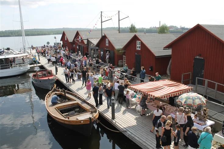 En liten marina vid Västra kajen camping med flera båtar förtöjda vid bryggorna i en lugn hamn, omgiven av vatten och grönska.