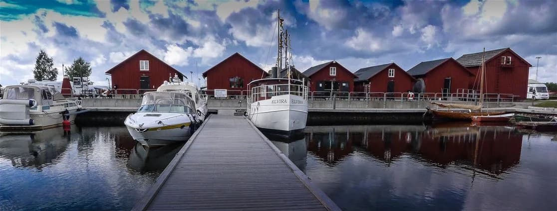 En rad av båtar förtöjda vid en marina på Västra Kajen Camping, spegelbilden av båtarna syns tydligt i det stilla vattnet. I bakgrunden kan man se fler båtar och bryggor som tillhör hamnen.
