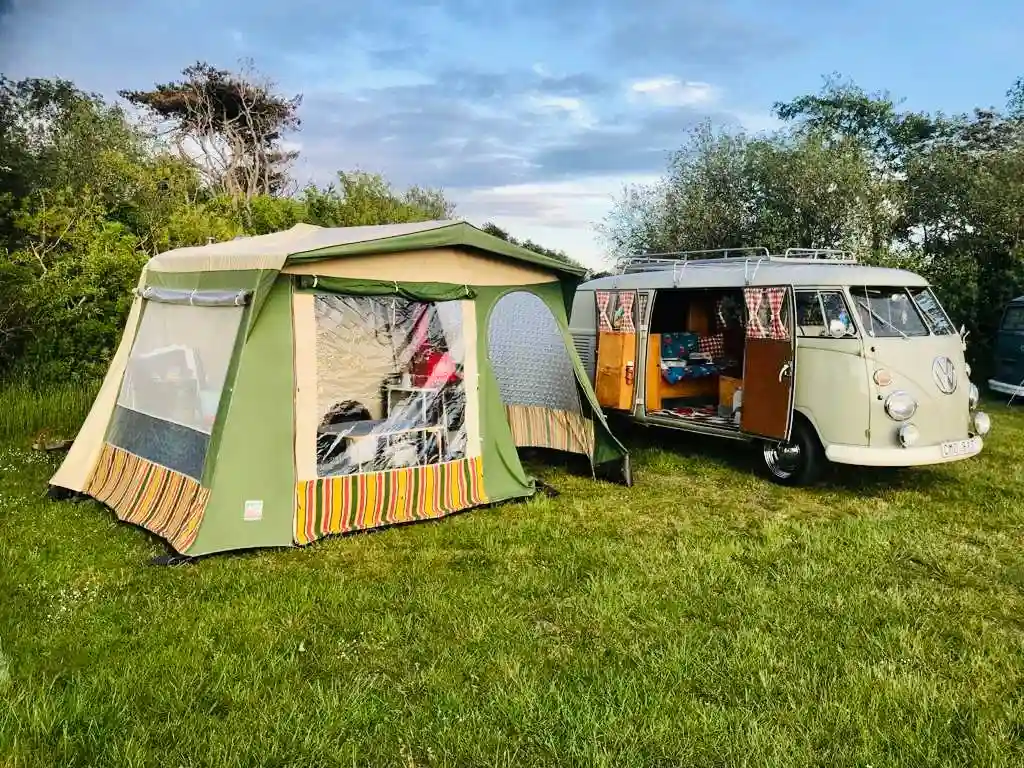 På bilden syns en campingplats med tält och skyddande presenningar. Det finns även en husbil i bakgrunden samt camping- och friluftsutrustning spridda runt omkring. Området är lugnt och avkopplande, perfekt för fritidsaktiviteter och rekreation.