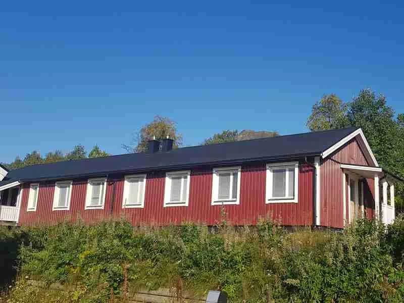 En lantlig stuga med röd fasad och vita knutar i Grönfjälls fritidsby, omgiven av en rymlig tomt och grönska, med mindre uthus i närheten. I bakgrunden kan man ana ett naturskönt landskap.