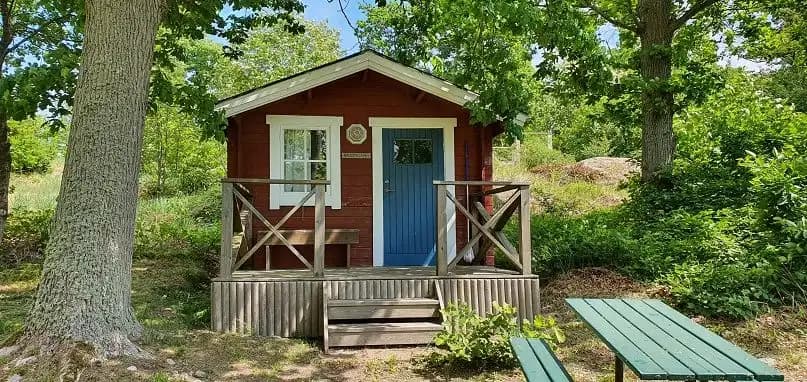 En liten trästuga i en lantlig miljö, omgiven av en grönskande trädgård på våren.