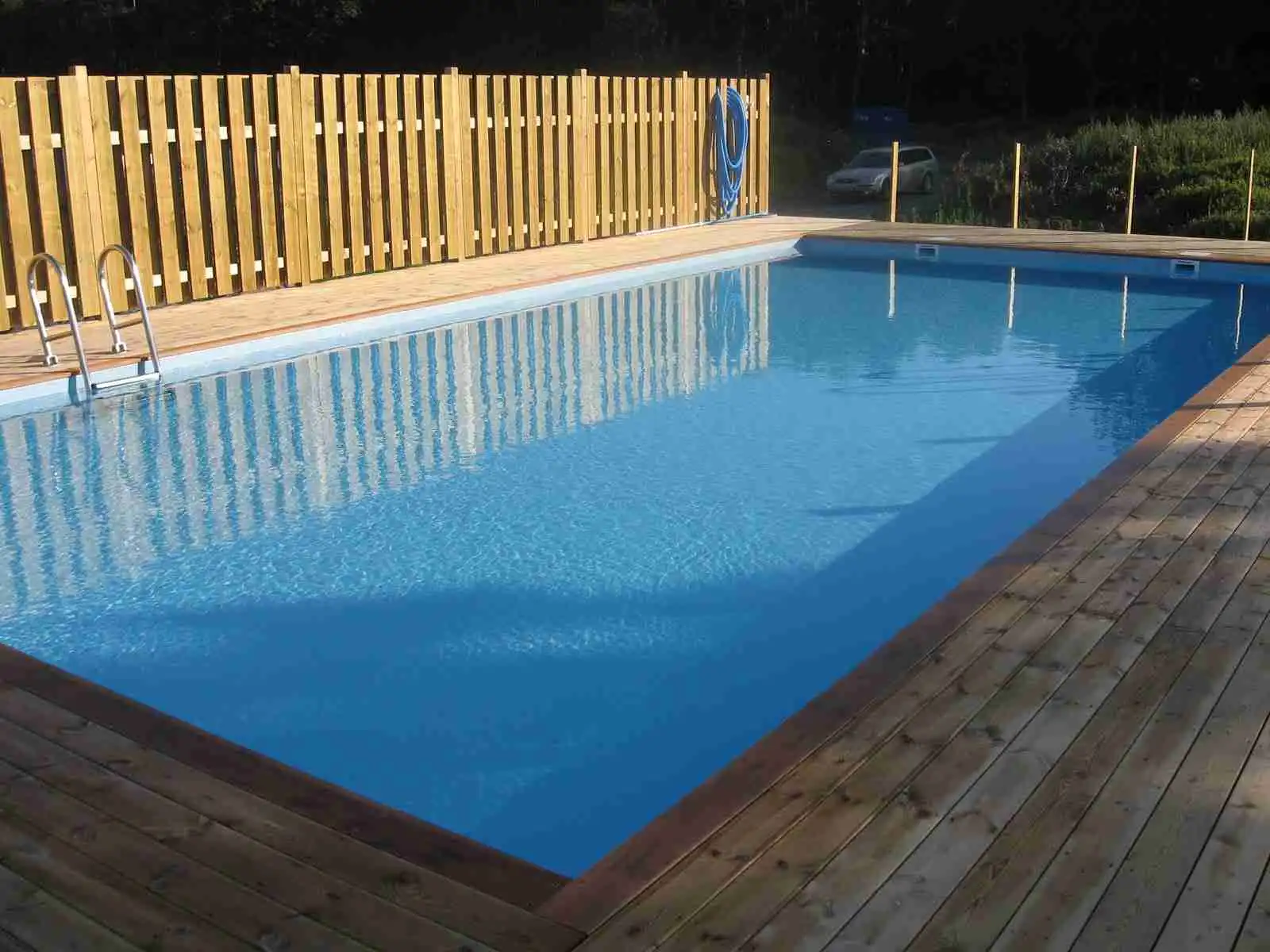 En solig dag vid Stråvalla Camping med en inbjudande pool omgiven av trästaket. Skuggiga områden under en pergola, perfekt för avkoppling vid detta friluftsområde.