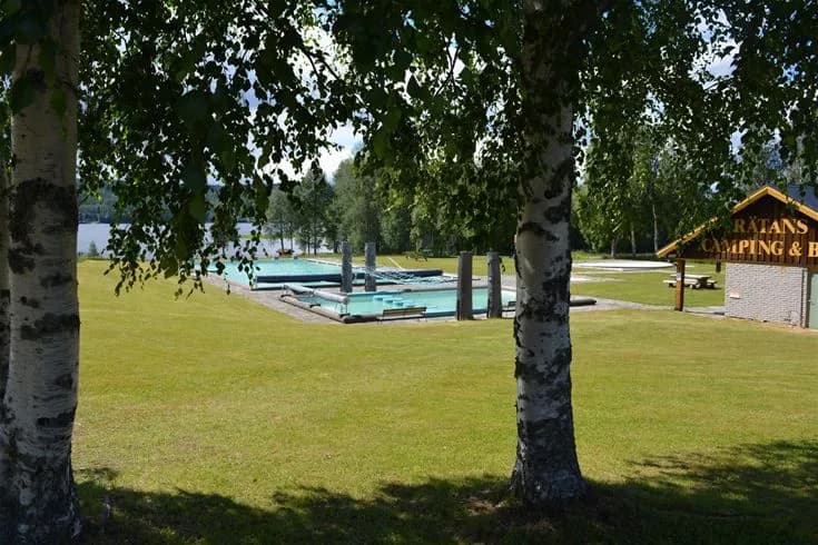 Bild av Rätans camping som visar grönskande träd och en välskött gräsmatta under en klar himmel. En gångväg leder genom området, och i bakgrunden syns en spegelblank swimmingpool som reflekterar omgivningen.
