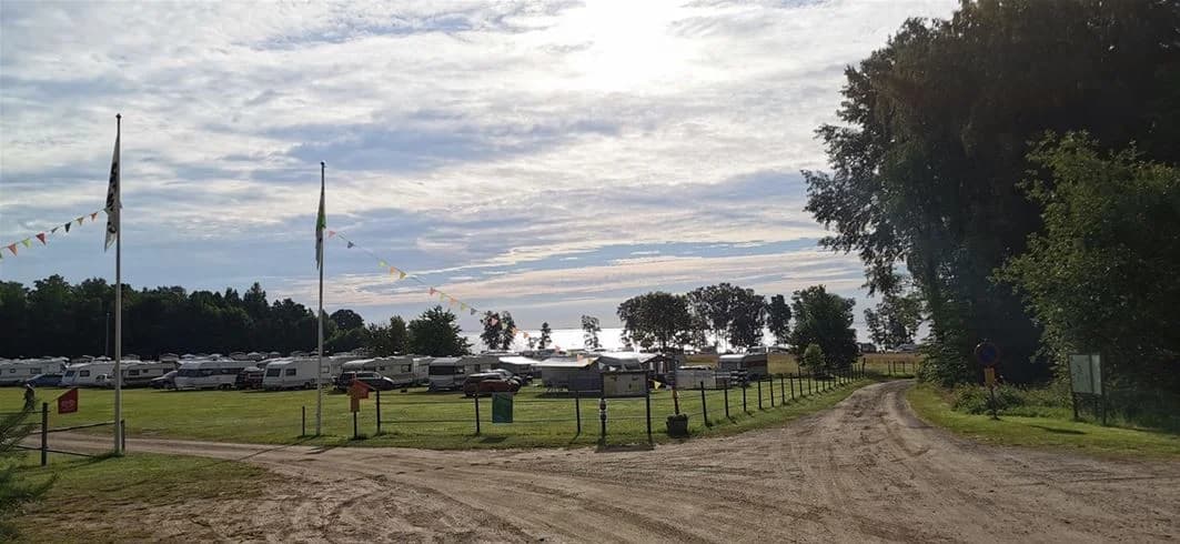 En campingplats med flera stora fordon och husbilar parkerade intill varandra på en grusad parkeringsyta. I bakgrunden syns träd och en öppen gräsyta.