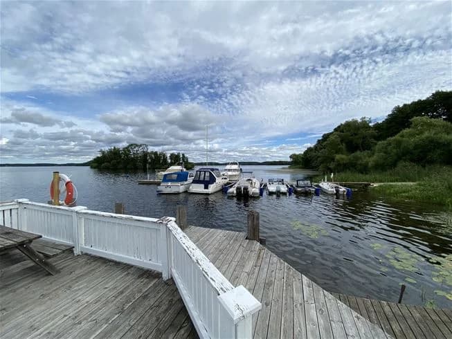 En campingplats vid en sjö med molnig himmel, båtar på vattnet och skogsklädda stränder i bakgrunden.
