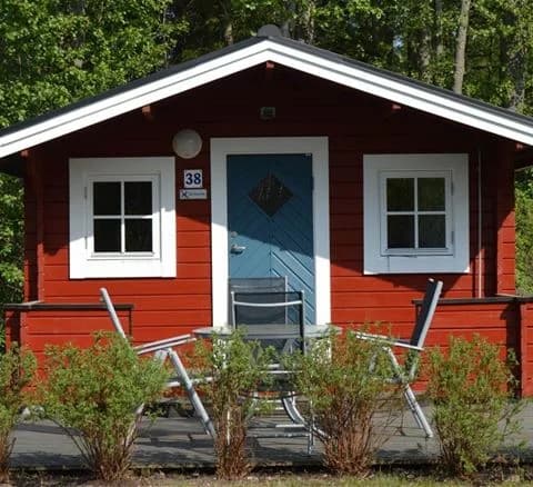 Bild av en rustik stuga vid Bödagårdens camping, omgiven av trä och grönska, med en träig fasad och enkel dörr.
