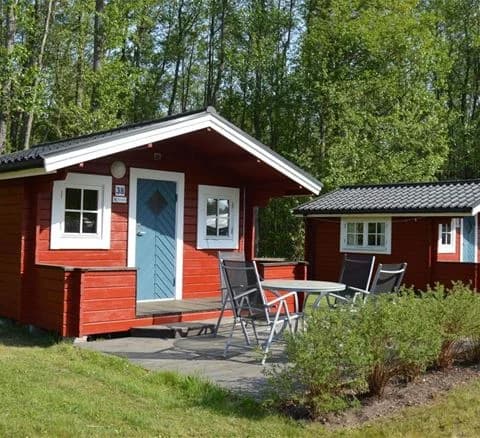 Bild av en mysig campingstuga omgiven av grönska på Bödagårdens camping. Stugan ligger i närheten av en välskött gräsmatta och trädgårdsbyggnader, vilket skapar en hemtrevlig och naturnära atmosfär.
