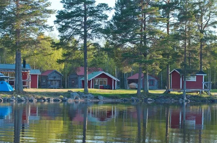 En pittoresk stuga vid strandkanten av en sjö. Stugan speglas vackert i det lugna vattnet, omgiven av grönska och kustlandskap. Perfekt för en fridfull naturupplevelse på Camp Viking.