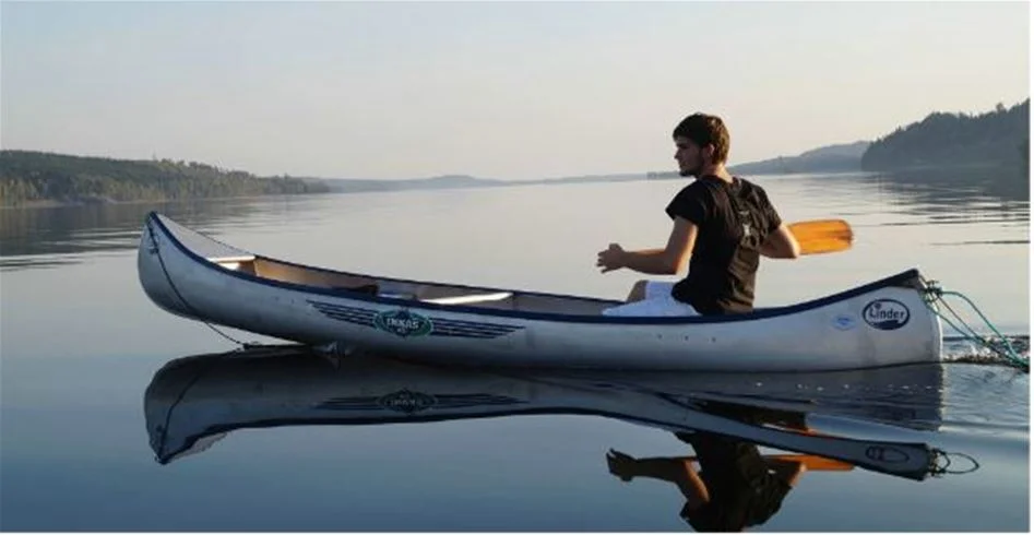 En man paddlar en kanot på en stilla sjö nära skotteksgården camping & stugby, omgiven av grönskande natur och klarblå himmel.