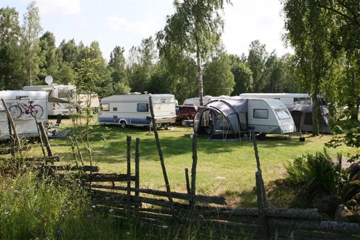En campingplats i Vimmerby med gröna ängar och träd, samt flera husvagnar och tält uppställda. En lantlig och avslappnande miljö perfekt för friluftsliv och rekreation.