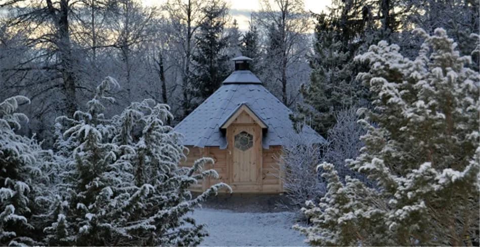 En snötäckt stuga omgiven av vinterlandskap, med frostiga grenar och en sluttning i bakgrunden, vid Åre camping.