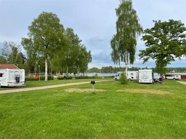 En campingplats med flera husbilar och husvagnar uppställda på en grön gräsmatta. Träden omger området och några besökare syns i närheten av fordonen.