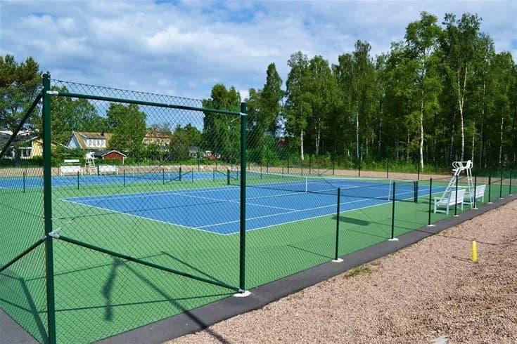 En grön tennisplan med omgivande stängsel på Gökaskratts camping. Planen är utrustad med ett nät i mitten och omges av grönt gräs för friluftsaktiviteter.