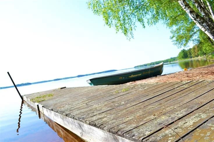 En träbrygga vid vattnet, med en liten båt förtöjd. Somrig atmosfär vid en sjö omgiven av trädkantade stränder.