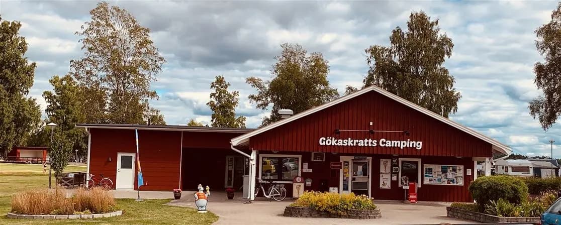 En livlig campingplats med besökare, sittplatser och bord utomhus, omgiven av grönskande natur och solsken. En restaurang och rekreationsområden syns i bakgrunden.