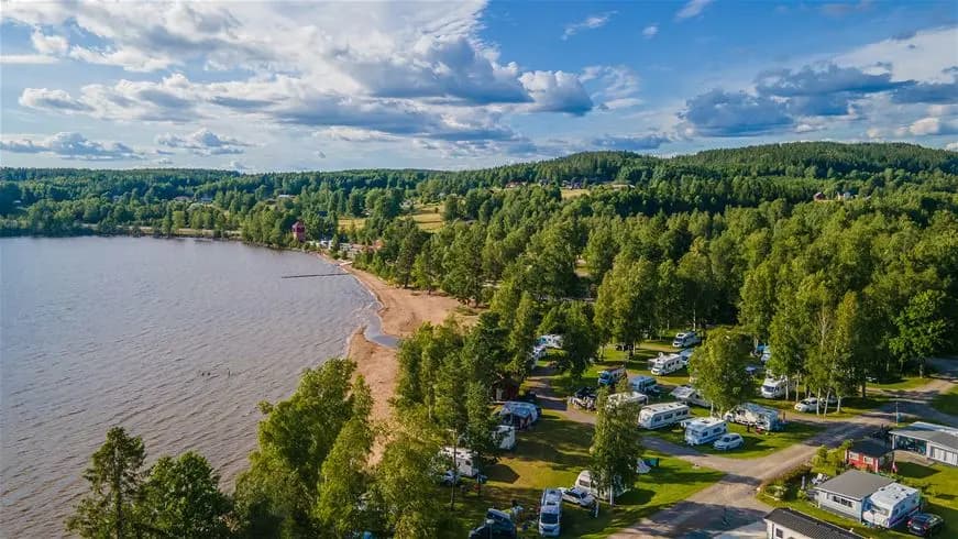 En naturskön campingplats vid vattnet med träd längs stranden och en sjö i bakgrunden. Restaurangens byggnad anas bland grönskan, vilket skapar en inbjudande atmosfär på Sandaholm Restaurang & Camping.