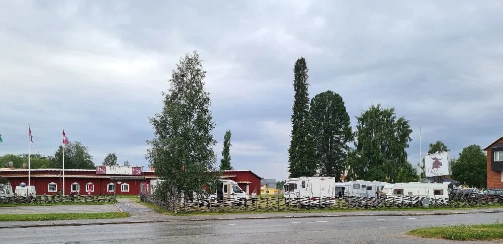 En stor asfaltparkering vid Hotel Nordica med flera parkerade bilar och husvagnar, omgiven av träd och en blå himmel.