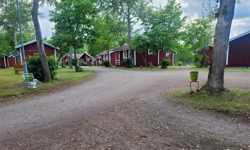 En pittoresk stuga omgiven av lummiga träd och välskött trädgård på Älvkarleby Fiskecamping. I förgrunden syns en grusad uppfart och buskar som ramar in den natursköna miljön.