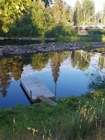 En pittoresk scen vid Ammeråns fiskecamp med en stilla vattendrag som slingrar sig genom det gröna landskapet. En bro sträcker sig över vattnet och speglas vackert i vattenytan, omgiven av frodig natur.