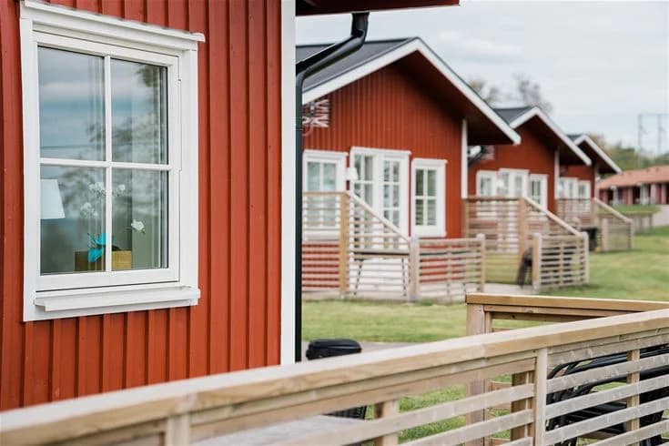 Bild av en mysig stuga med träpanel på Mösseberg camping. Stugan har en veranda med trätrall och utemöbler, omgiven av skuggiga grönområden.