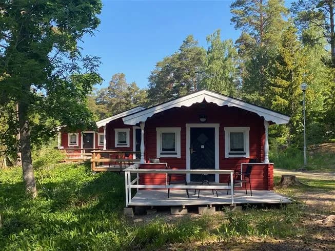 En mysig timmerstuga på Ängskärs havscamping, omgiven av en lummig trädgård med grönskande gräsmatta och skuggiga trädkronor, inbjudande veranda i förgrunden.
