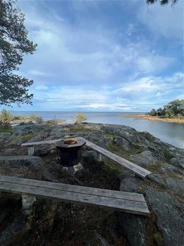 Vy över ängskärs havscamping med kustlandskap, där en sandig strand möter det lugna skärgårdsvattnet. I bakgrunden syns grönskande träd och en blå himmel.