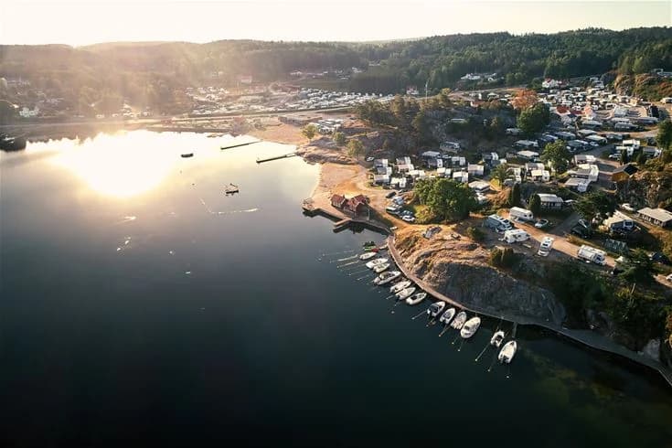 En lugn vattenlagun med flera små båtar förtöjda vid en brygga, omgiven av grönskande kustlandskap och ett campingområde i bakgrunden.